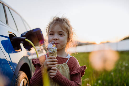 Ein junges Mädchen genießt ein Eis, während ihre Familie ihr Elektrofahrzeug auflädt. - HPIF31871