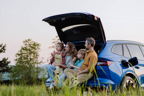 Eine fröhliche Familie wartet darauf, dass ihr Auto wieder aufgeladen wird, während sie sich auf ein Picknick vorbereitet, bequem im Kofferraum sitzend - HPIF31873