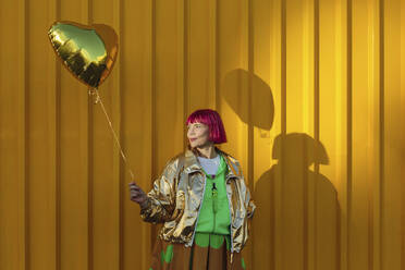 Lächelnde ältere Frau, die einen herzförmigen Ballon vor einer gelben Wand hält - LLUF01128