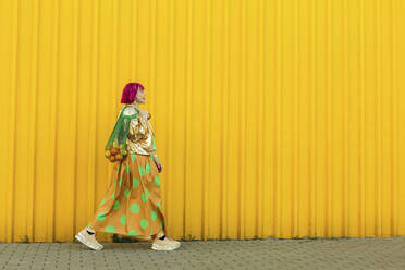 Senior woman carrying fruits in mesh bag and walking near yellow wall - LLUF01134