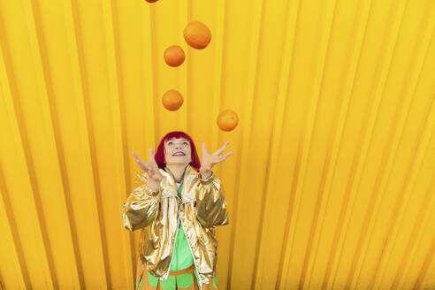 Glückliche ältere Frau, die vor einer gelben Wand mit Orangen jongliert - LLUF01136