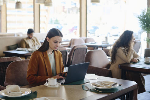 Junge Geschäftsfrau sitzt mit Kaffeetasse und arbeitet am Laptop im Restaurant - DSHF01396