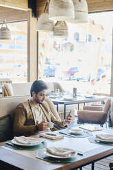 Junger Geschäftsmann hält eine Kaffeetasse und benutzt sein Smartphone im Restaurant - DSHF01398