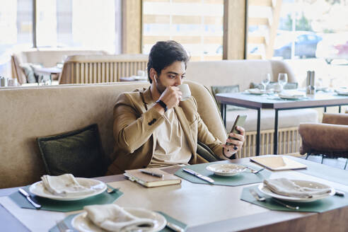 Junger Geschäftsmann trinkt Kaffee und benutzt sein Smartphone im Restaurant - DSHF01399