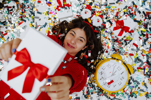 Lächelndes Mädchen hält Geschenkbox und liegt auf Konfetti in der Nähe der Uhr - MDOF01738