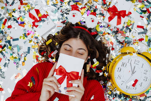 Mädchen versteckt Gesicht mit Geschenkbox und liegt auf Konfetti in der Nähe der Uhr - MDOF01739
