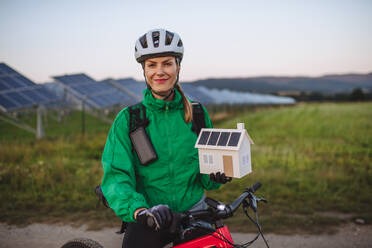 Porträt einer schönen Radfahrerin, die während einer sommerlichen Radtour in der Natur vor Solarmodulen in einem Solarpark steht. Radfahrer hält ein Modell eines Hauses mit Photovoltaik-Modulen auf dem Dach. Solarenergie - nachhaltige Energiezukunft. - HPIF31883