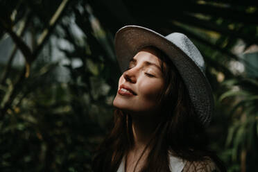 Portrait einer jungen Frau mit Hut und geschlossenen Augen im botanischen Garten. Schöne Botanikerin im Dschungel, Seitenansicht. - HPIF31885