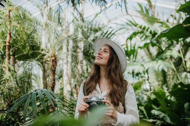 Porträt einer jungen Fotografin mit Hut und Rucksack im botanischen Garten. Botanikerin im Gewächshaus beim Fotografieren. - HPIF31887