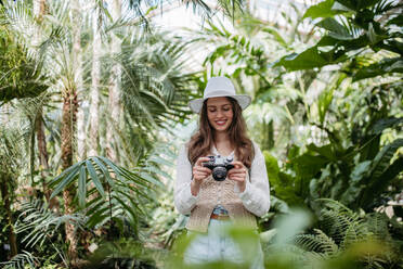 Porträt einer jungen Fotografin mit Hut und Rucksack im botanischen Garten. Botanikerin im Gewächshaus beim Fotografieren. - HPIF31888