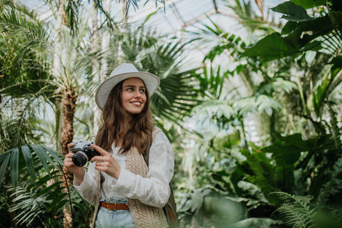 Porträt einer jungen Fotografin mit Hut und Rucksack im botanischen Garten. Botanikerin im Gewächshaus beim Fotografieren. - HPIF31889