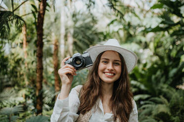 Porträt einer jungen Fotografin mit Hut und Rucksack im botanischen Garten. Botanikerin im Gewächshaus beim Fotografieren. - HPIF31892