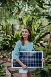 Junge Frau im Gewächshaus mit Modell eines Hauses mit Sonnenkollektoren, Konzept der erneuerbaren Energien und des Naturschutzes. - HPIF31898