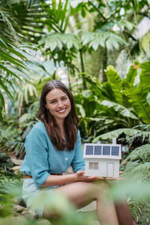 Junge Frau im Dschungel, die ein Modell eines Hauses mit Sonnenkollektoren hält, Konzept der erneuerbaren Energien und des Naturschutzes. - HPIF31905