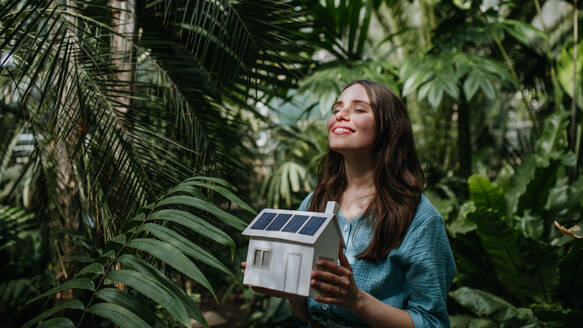 Junge Frau im Dschungel, die ein Modell eines Hauses mit Sonnenkollektoren hält, Konzept der erneuerbaren Energien und des Naturschutzes. - HPIF31906