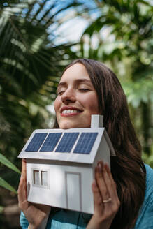 Junge Frau im Dschungel, die ein Modell eines Hauses mit Sonnenkollektoren hält, Konzept der erneuerbaren Energien und des Naturschutzes. - HPIF31907
