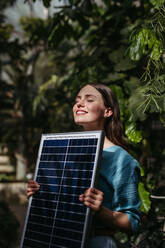 Junge Frau im Gewächshaus mit Modell eines Hauses mit Sonnenkollektoren, Konzept der erneuerbaren Energien und des Naturschutzes. - HPIF31909