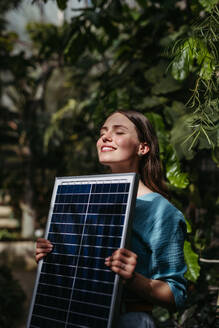 Junge Frau im Gewächshaus mit Modell eines Hauses mit Sonnenkollektoren, Konzept der erneuerbaren Energien und des Naturschutzes. - HPIF31909