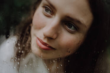 Portrait of a young woman in misty greenhouse through foggy glass with water droplets. - HPIF31913