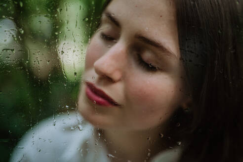 Porträt einer jungen Frau in einem nebligen Gewächshaus durch ein nebliges Glas mit Wassertropfen. - HPIF31915