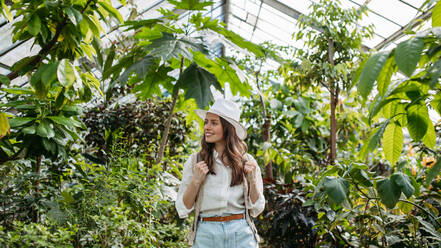 Porträt einer jungen Frau mit Hut und Rucksack im botanischen Garten, Botanikerin im Gewächshaus. - HPIF31932