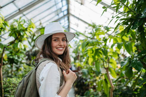 Porträt einer jungen Frau mit Hut und Rucksack im botanischen Garten, Botanikerin im Gewächshaus. - HPIF31939