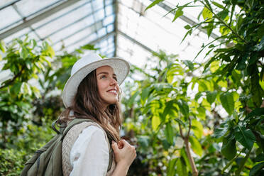 Porträt einer jungen Frau mit Hut und Rucksack im botanischen Garten, Botanikerin im Gewächshaus. - HPIF31940