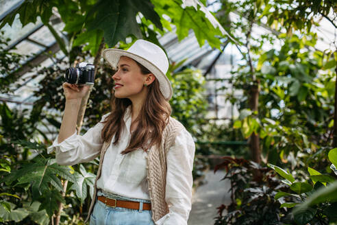 Porträt einer jungen Fotografin mit Hut und Rucksack im botanischen Garten. Botanikerin im Gewächshaus beim Fotografieren. - HPIF31943