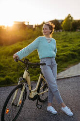 Young woman standing by electro bicycle, during sunset, sunrise. Concept of commuting and ecologic traveling. - HPIF31990