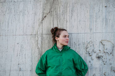 Waist up portrait of young city woman in green windbreaker jacket leaning against a concrete wall during sunset. Copy space image. - HPIF31999