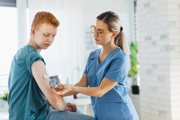 Diabetologist doctor connecting continuous glucose monitor sensor to smartphone, checking teenage boy's glucose data in the real time. Endocrinologist taking care of teenage diabetic patient. - HPIF32139
