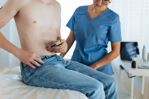 Ein Diabetologe zeigt einem jugendlichen Patienten, wie man Insulin in den Bauch spritzt, indem er einen Insulin-Pen benutzt. Ein Endokrinologe spritzt Medizin in den Bauch eines Jungen. - HPIF32145