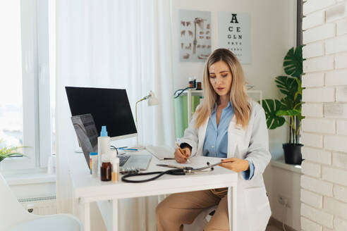 Young beautiful female doctor working on laptop in doctor's office. Physician doing paperwork and administrative tasks. - HPIF32153