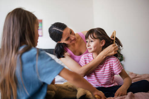 Porträt einer Mutter mit zwei Töchtern auf einem Bett im Schlafzimmer. Die Mutter kämmt die langen Haare ihrer Tochter mit einem Kamm. Konzept des Muttertags und der mütterlichen Liebe. - HPIF32163