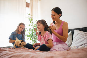 Porträt einer Mutter mit zwei Töchtern auf einem Bett im Schlafzimmer. Die Mutter kämmt die langen Haare ihrer Tochter mit einem Kamm. Konzept des Muttertags und der mütterlichen Liebe. - HPIF32164