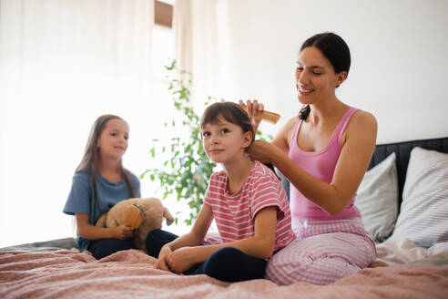 Porträt einer Mutter mit zwei Töchtern auf einem Bett im Schlafzimmer. Die Mutter kämmt die langen Haare ihrer Tochter mit einem Kamm. Konzept des Muttertags und der mütterlichen Liebe. - HPIF32165