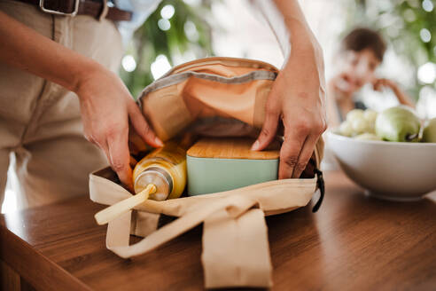 Mutter packt Schultasche für ihr Kind. Sie packt eine Flasche Saft und Snacks für ihre Kinder in die Schultasche. Snack, Schulessen in einer Snackbox für die Schule. - HPIF32183