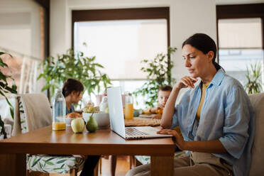 Mutter, die am Küchentisch am Laptop arbeitet, während die Kinder frühstücken. Fernarbeit und Home Office für Mütter mit Kindern. - HPIF32185