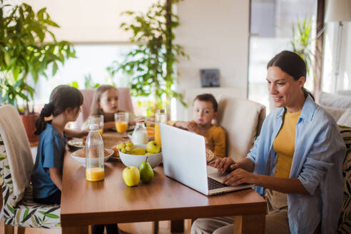 Mutter, die am Küchentisch am Laptop arbeitet, während die Kinder frühstücken. Fernarbeit und Home Office für Mütter mit Kindern. - HPIF32186