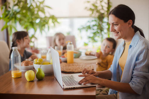 Mutter, die am Küchentisch am Laptop arbeitet, während die Kinder frühstücken. Fernarbeit und Home Office für Mütter mit Kindern. - HPIF32187