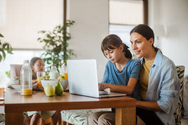 Mutter, die am Küchentisch am Laptop arbeitet, während die Kinder frühstücken. Fernarbeit und Home Office für Mütter mit Kindern. - HPIF32189