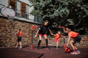 Mom playing football with her children, dressed in football jerseys. The family as one soccer team. Fun family sports activities outside in the backyard or on the street. - HPIF32194