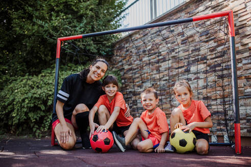 Mom playing football with her children, dressed in football jerseys. The family as one soccer team. Fun family sports activities outside in the backyard or on the street. - HPIF32195