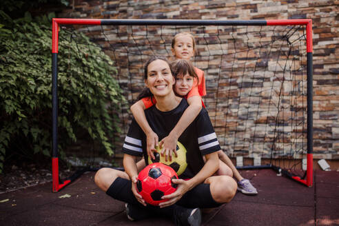 Mom playing football with her daughters, dressed in football jerseys. The family as one soccer team. Fun family sports activities outside in the backyard or on the street. - HPIF32198