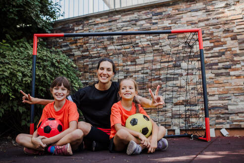 Mom playing football with her daughters, dressed in football jerseys. The family as one soccer team. Fun family sports activities outside in the backyard or on the street. - HPIF32199