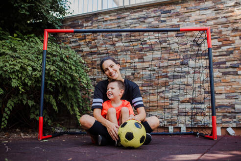 Mom playing football with her son, dressed in football jerseys. The family as one soccer team. Fun family sports activities outside in the backyard or on the street. - HPIF32200