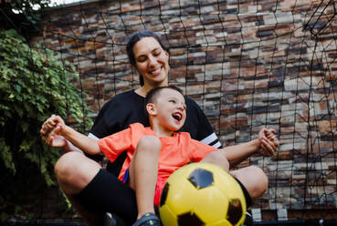 Mom playing football with her son, dressed in football jerseys. The family as one soccer team. Fun family sports activities outside in the backyard or on the street. - HPIF32201