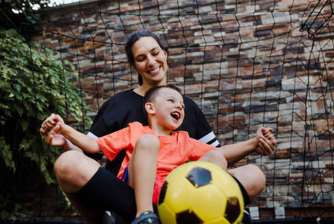 Mom playing football with her son, dressed in football jerseys. The family as one soccer team. Fun family sports activities outside in the backyard or on the street. - HPIF32201
