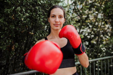 Portrait of confident boxer woman in red boxing gloves standing outdoors. Woman boxing for relaxation and bolsting mental health. Boxing empowers women. - HPIF32203