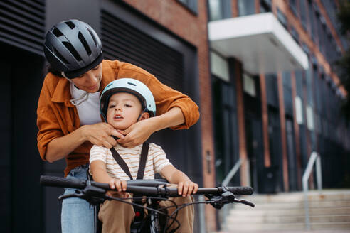 Mutter, die ihrem Sohn den Fahrradhelm auf den Kopf setzt und ihn auf dem Kinderfahrradträger oder -sitz trägt. Mutter, die mit einem kleinen Kind mit dem Fahrrad durch die Stadt fährt. - HPIF32204